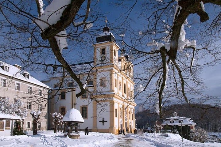Maria Plain pilgrimage church viewed from beneath the iconic tree, surrounded by a snowy winter landscape near Salzburg.